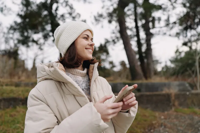 Woman in a warm winter coat and beanie smiles while using a phone outdoors in a park, capturing a candid moment of everyday technology and nature around her