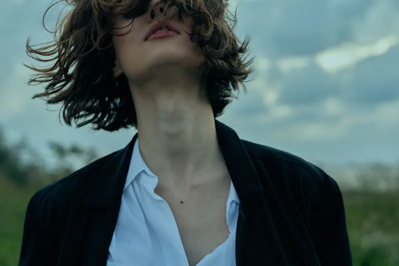 Young adult female in a casual blazer and white shirt stands in a breezy outdoor fashion setting, windblown hair and confident posture, moody atmosphere for lifestyle fashion.