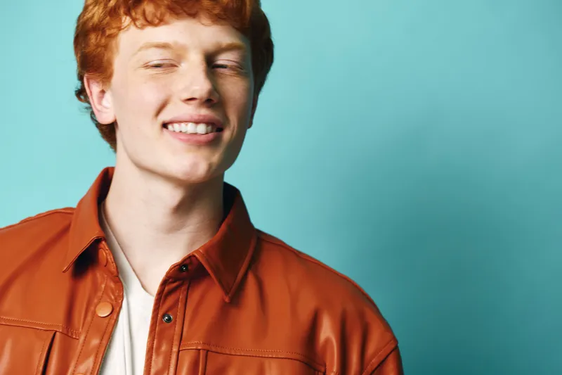Young man with red hair smiling confidently in orange leather jacket against turquoise background. Casual style, light skin, relaxed pose and joyful expression in studio portrait.
