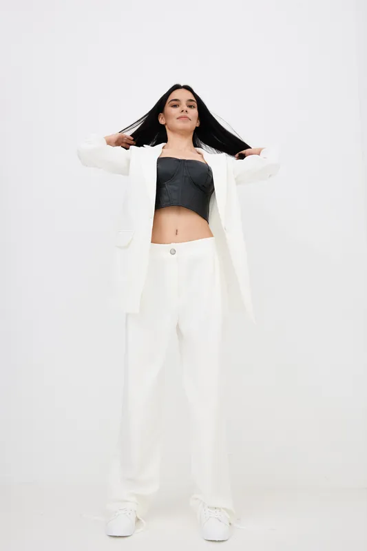 confident woman with long black hair wearing white pants and jacket, posing in modern minimalistic style against plain white background, casual fashion look