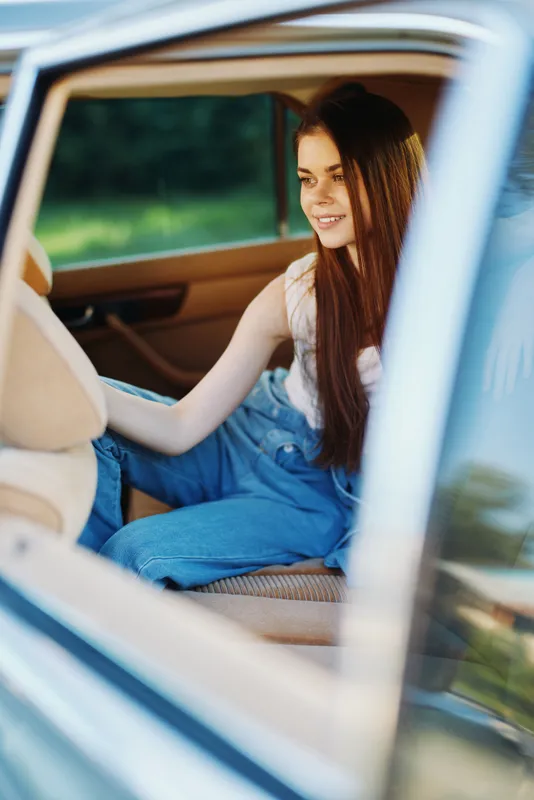 Happy young woman sitting in a car, smiling joyfully, wearing casual denim overalls, outdoors in a natural green background.