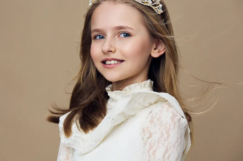 Portrait of a young girl in a white lace dress and crown, exuding joy and elegance with a soft beige background, perfect for children s fashion and celebrations