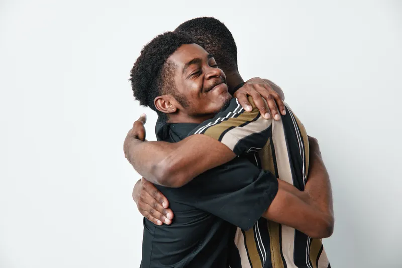 Two young black men hugging each other with eyes closed and smiles, showing friendship and affection. Casual clothing and dark skin tones on white background.