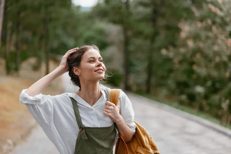 Young woman in casual attire enjoying nature, smiling confidently on a sunny day, wearing a light shirt and green overalls, perfect for promoting outdoor joy and lifestyle