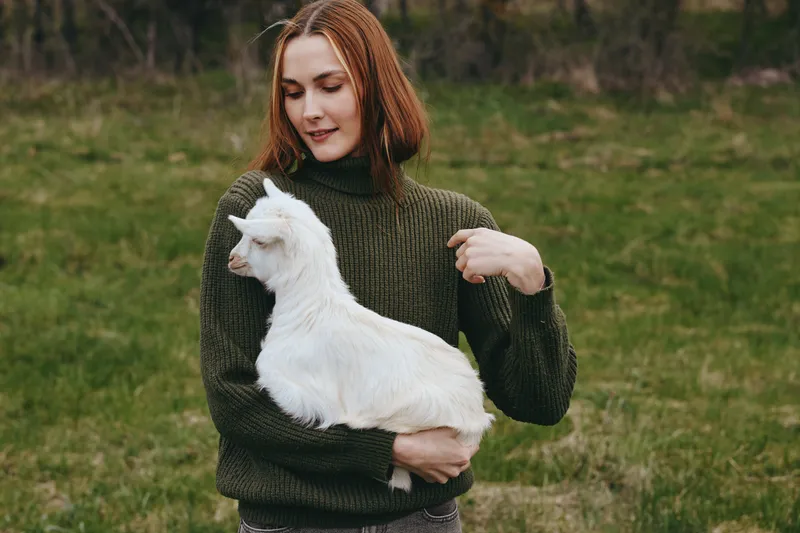 A woman in a dark green sweater holds a white goat kid in a grassy field, smiling softly. A candid lifestyle moment highlighting bonding with animals and rural simplicity.