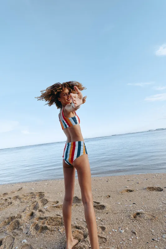 Beach, smiling girl, colorful swimsuit, standing by calm ocean, curly hair in sunlight, childhood summer vacation, clear sky, soft breeze, natural colors
