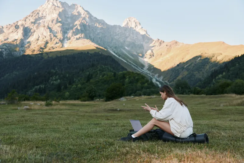 A cheerful professional sits outdoors in a grassy field with a laptop, smiling and focused, enjoying a calm and productive work moment amid nature.