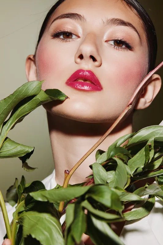 Close-up portrait of a woman with shiny red lips and glowing skin surrounded by fresh green leaves. Creative beauty fashion concept with a natural and elegant mood in the studio.