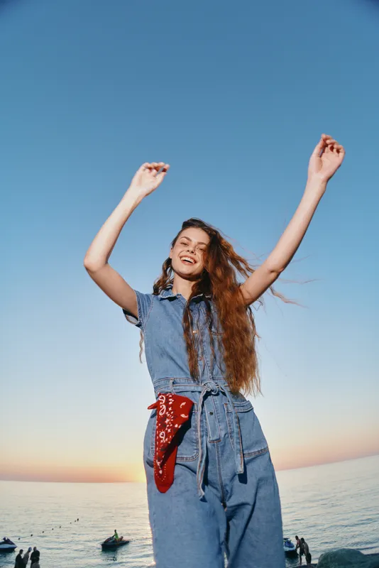 Lifestyle woman in modern boho-western denim street style joyfully raising arms by the sea during sunset with a film color aesthetic.