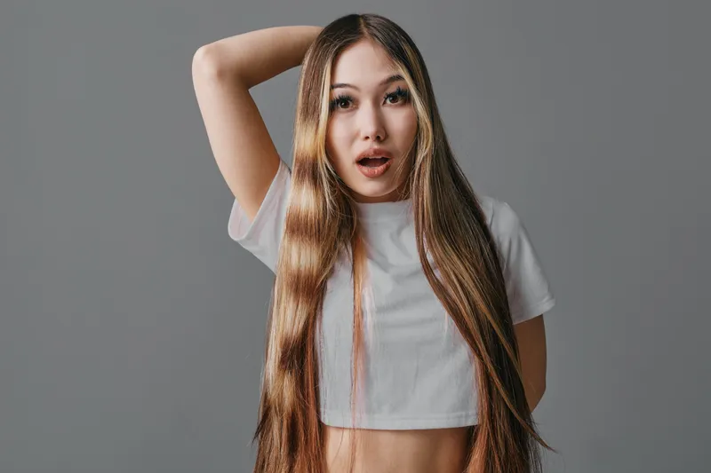Young woman with long straight hair and surprised facial expression wearing white crop top and posing with hand behind head on gray background studio.