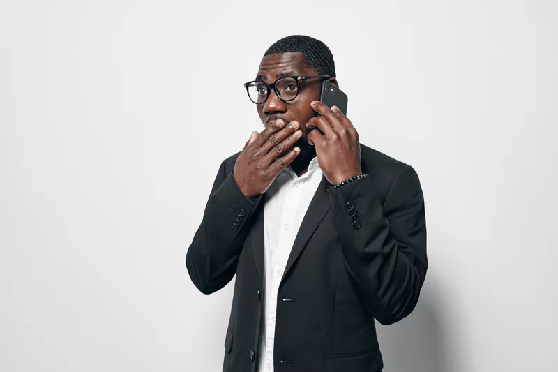 African American man with dark skin wearing glasses and black suit talking on smartphone with surprised expression on white background. Business communication and technology concept.