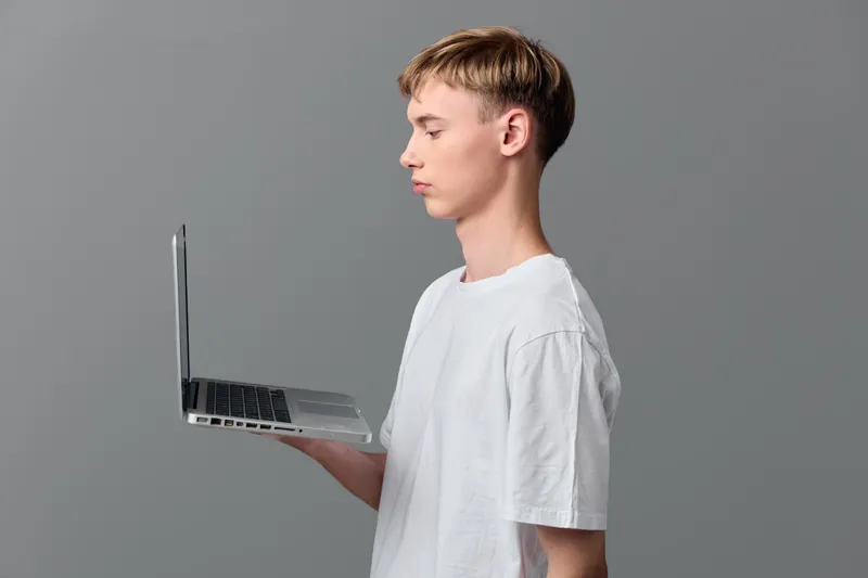 Young man in a white t shirt holds a laptop in side view, showcasing technology and portable computing for work, study, or creative tasks in a clean studio setup