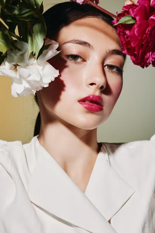 Portrait of young woman with fresh natural makeup wears white clothing and vibrant flowers in hair, highlighting clean skin and bright pink lips. Beauty fashion creative concept.