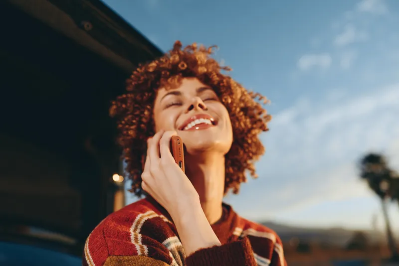 Woman lifestyle, smartphone calling outdoors in daylight. Curly haired smiling woman in striped sweater holds phone to ear, relaxed candid moment with soft blue sky background.