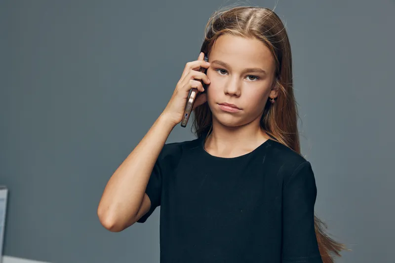 Girl child talking on smartphone in room wearing black top looking serious with long hair. Communication, technology and youth with person holding phone for conversation or call.