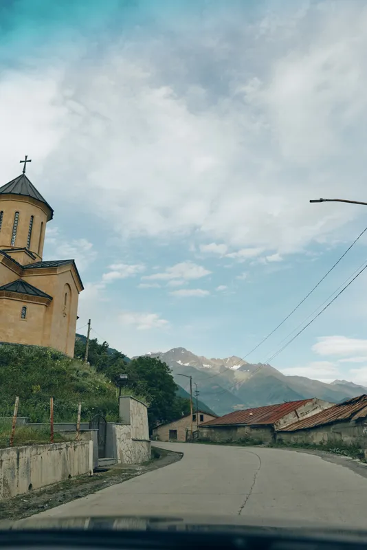 Rural road with a small church on the roadside, mountain backdrop and blue sky, capturing a peaceful travel moment through a quaint village setting