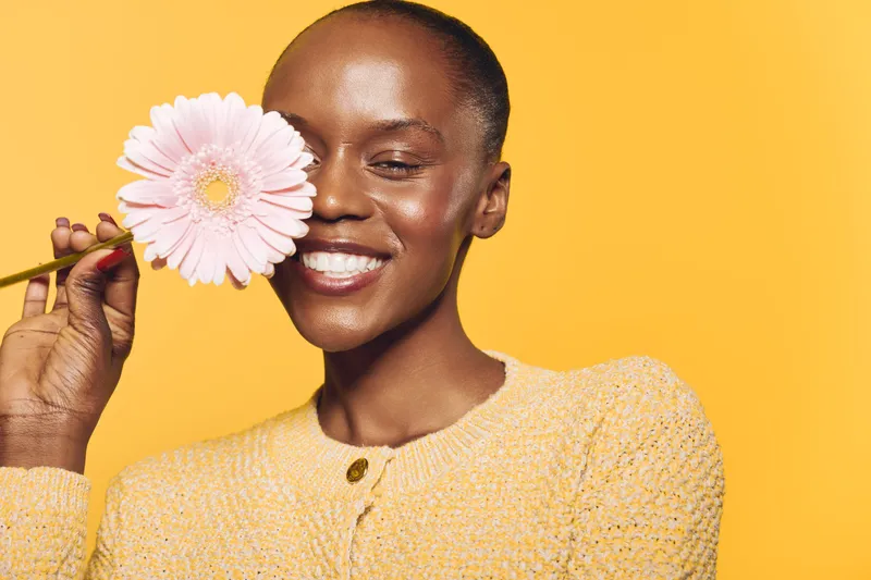 Smiling black woman with dark skin holds pink flower near face, wearing yellow sweater. Bright yellow background creates cheerful mood. Concept for beauty, happiness, and natural skincare.