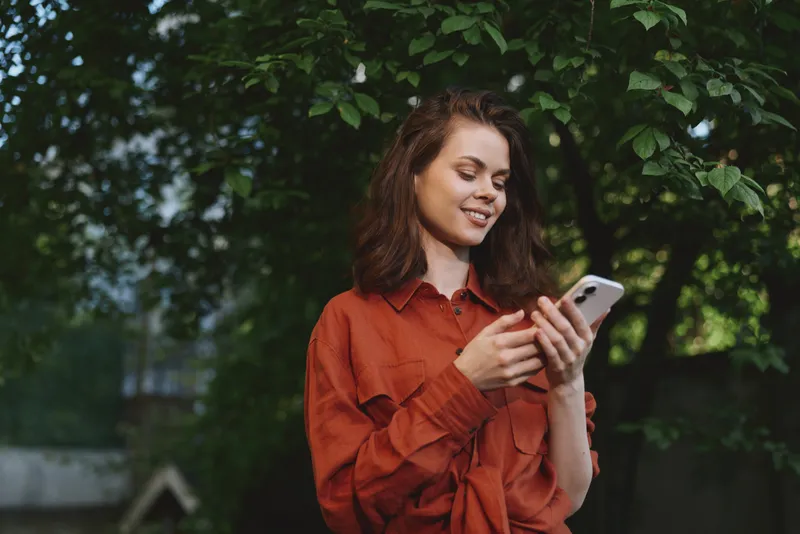 Casual young woman in an orange shirt stands outdoors among lush green trees, checking her phone with a gentle smile, creating a relaxed, natural mood while sun dappled leaves create a warm, inviting