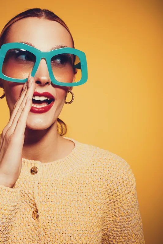 Young woman with large turquoise sunglasses and red lipstick whispers with hand near mouth on yellow background. Concept of secret, communication, and fashion style.