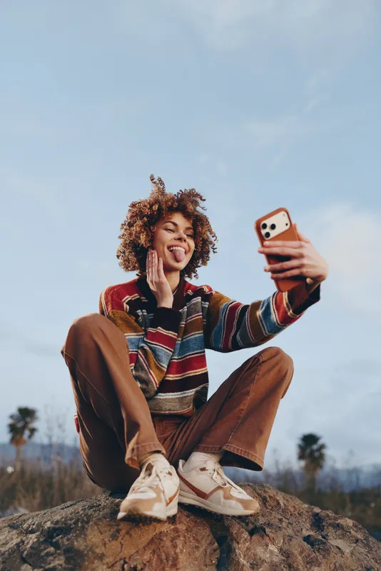 Woman with curly hair smiling selfie holding phone outdoors on a rocky hill, casual lifestyle portrait under blue sky daylight, warm relaxed mood, nature background