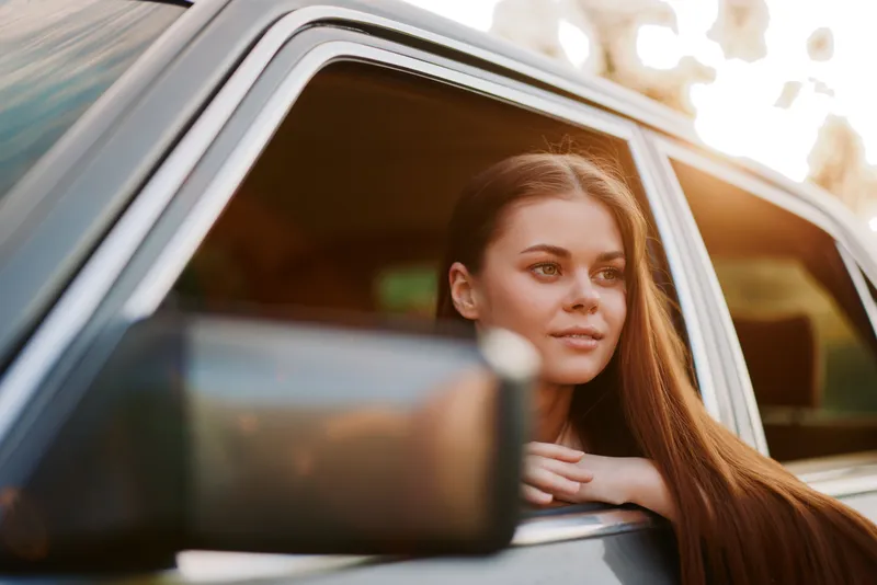 Young woman with long hair looking out of a car window, enjoying a sunny day, reflecting happiness and freedom in her expression and surroundings.