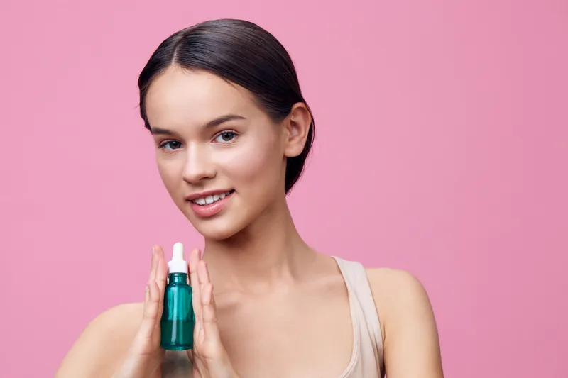 Young woman with natural makeup holding a green skincare serum on a pink background, showcasing healthy skin and beauty