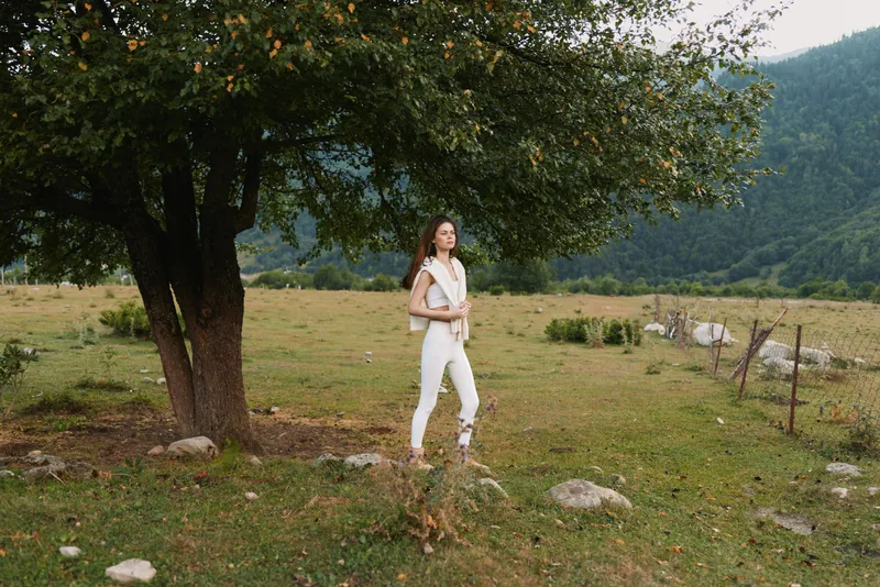 Young woman in white outfit stands near tree in green meadow with mountains in background, enjoying nature and peaceful outdoor lifestyle on sunny day.