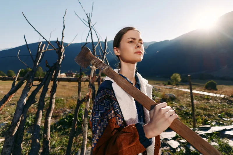 Woman in countryside field holding an axe over shoulder by wooden fence, mountains in background and sunlight. Rustic portrait of a young farmer in rural landscape and nature.
