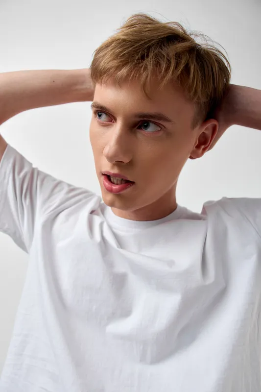 Young man stretching arms behind head in a casual white shirt, relaxed pose and thoughtful expression, studio lighting, simple background, lifestyle and wellness vibe