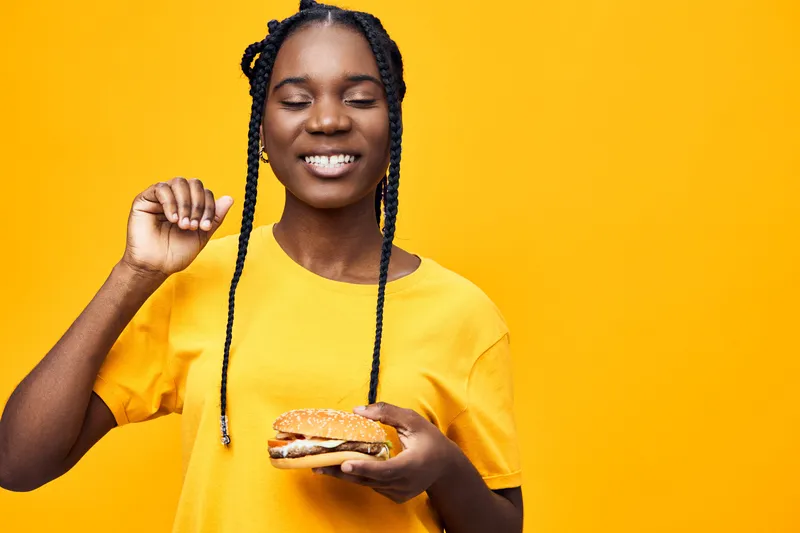 Happy young Black woman enjoying a delicious burger against a vibrant yellow background for healthy eating and food joy concept