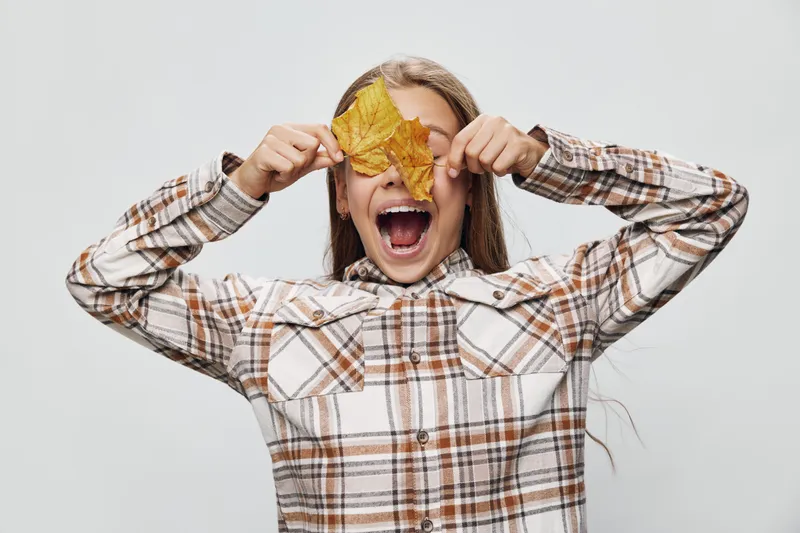 Girl with long hair wearing plaid shirt holds yellow autumn leaves over eyes with open mouth on white background for seasonal fun and fall nature concept.