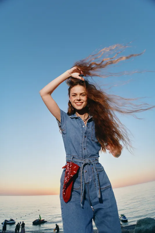 modern boho-western denim street style woman smiling with long hair outdoors by the sea at sunset, film color, carefree and vibrant lifestyle moment