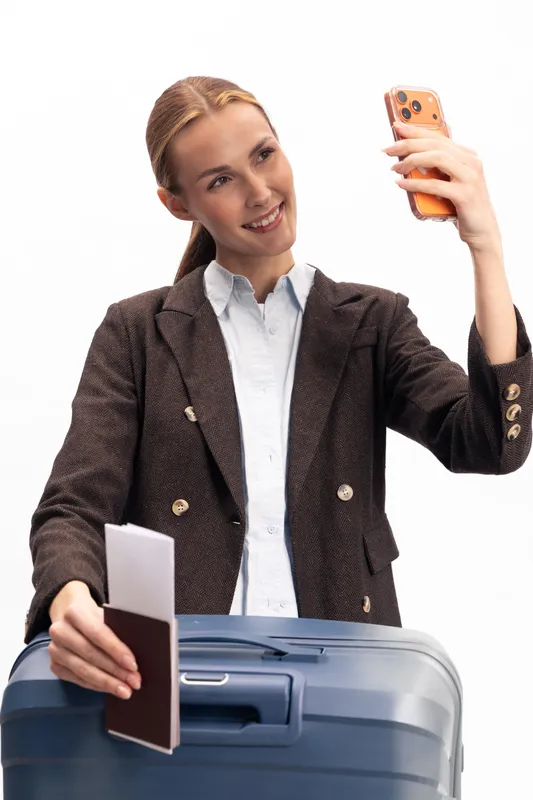 Young woman in brown blazer takes a selfie with smartphone while holding passport and standing behind blue suitcase. Concept for travel, vacation, tourism, and modern lifestyle.