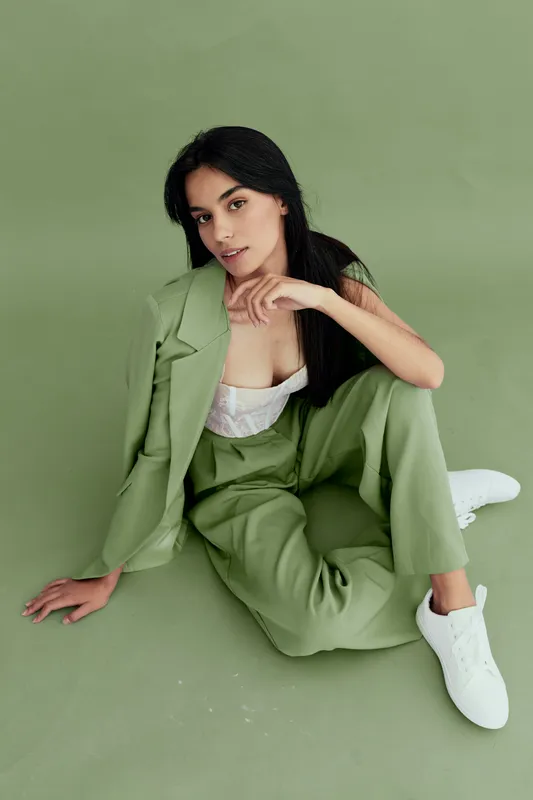 Young woman in green suit posing seated on floor with casual white sneakers against plain green background, showcasing modern fashion and relaxed style.