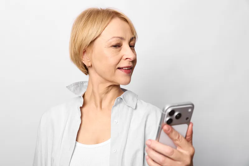Elderly woman in white blouse holds a smartphone, serious senior technology mood. She reads a message on the screen indoors against a clean white background, closeup calm.