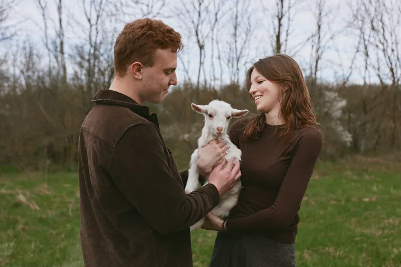 Young couple holding a baby goat outdoors in a grassy field during early spring, sharing a joyful moment with soft natural light and casual clothing.