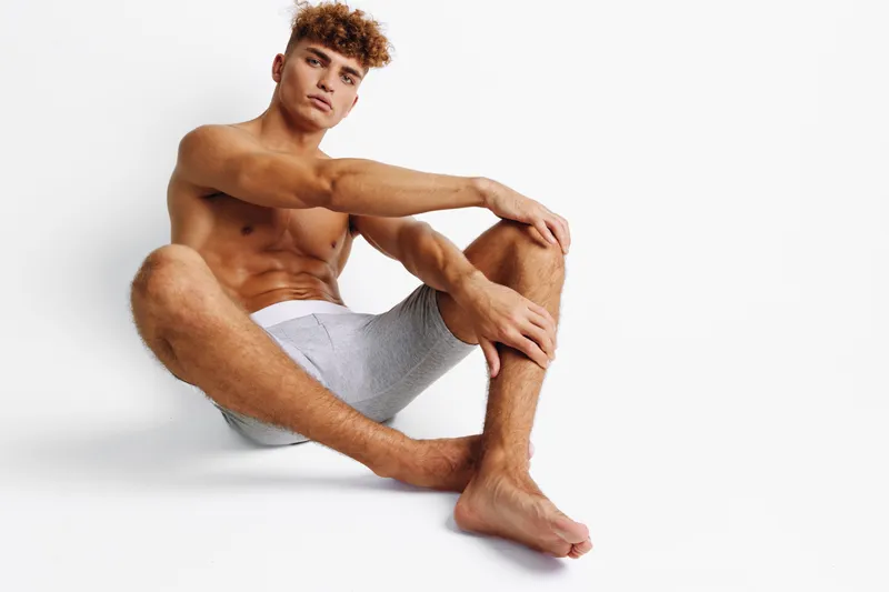 Young man with curly hair sitting on floor in underwear against white background, posing with relaxed expression and muscular body in studio lighting.