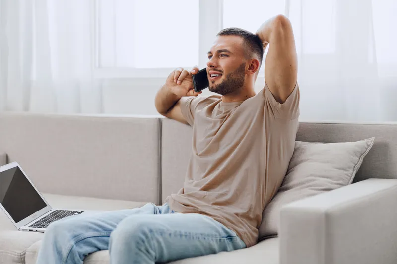 Relaxed man enjoying a phone call while sitting on a couch, exuding a sense of comfort and tranquility in a modern, bright living space.