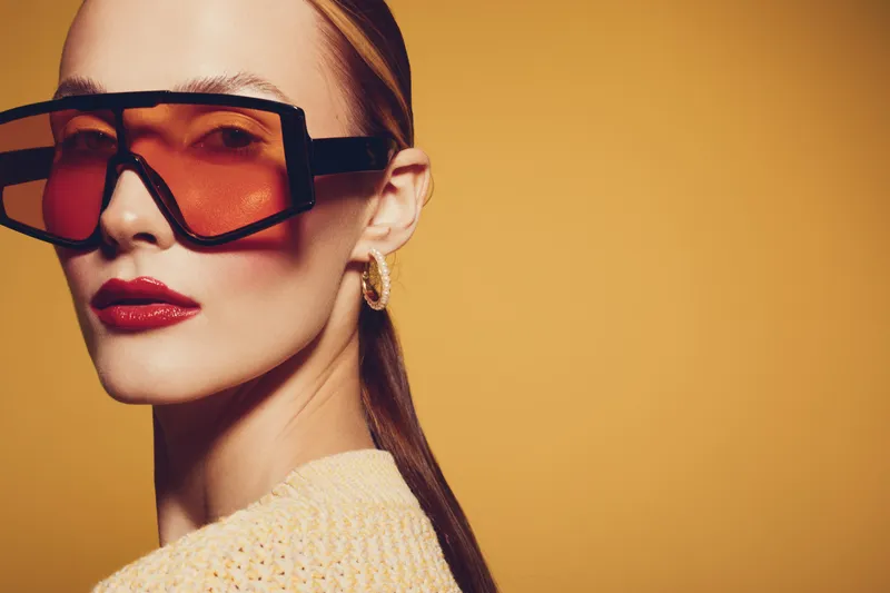 Stylish woman with slicked back hair wearing oversized orange sunglasses and red lipstick poses against a warm yellow background. Fashion concept with bold accessories and makeup.
