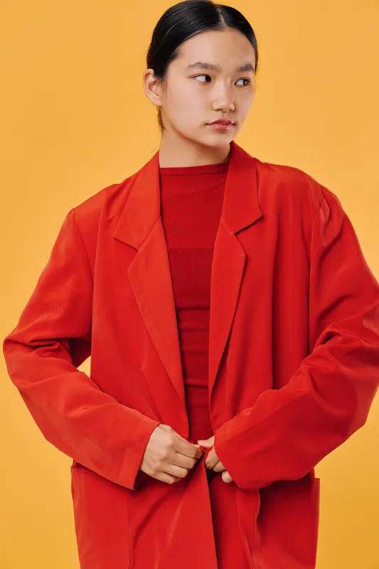 Young woman wearing oversized red jacket and red shirt posing on yellow background. Asian female model with serious expression and casual style in studio setting.