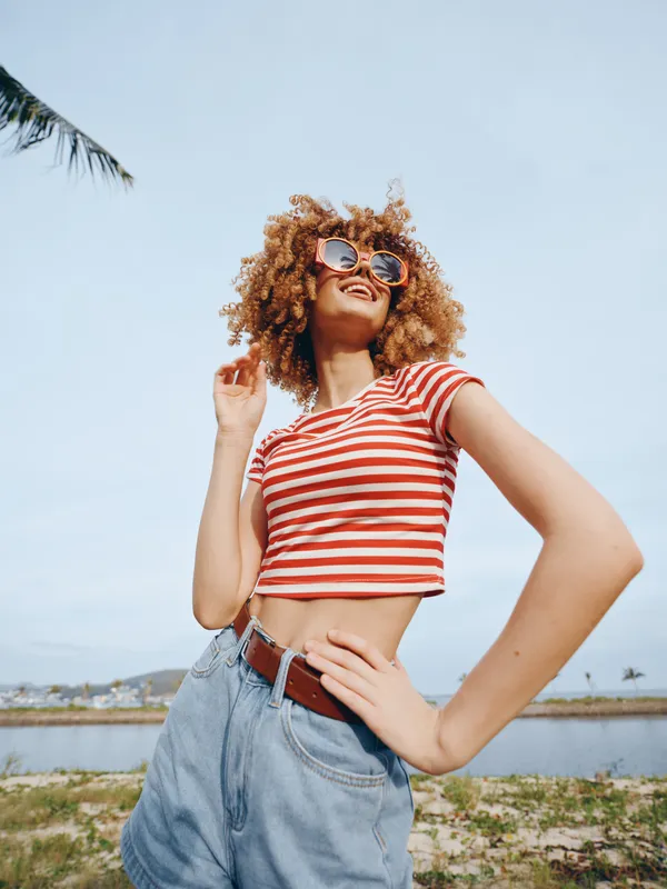 Stylish young woman posing outdoors in bright sunlight, wearing a red striped crop top, high waisted denim shorts, and sunglasses, exuding carefree summer energy in a landscape.