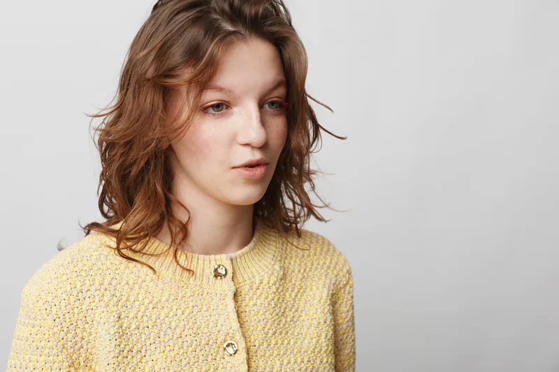 Young woman with wavy brown hair wearing a yellow knitted cardigan looks thoughtfully to the side on a plain light background. Concept of contemplation, casual style, and natural beauty.