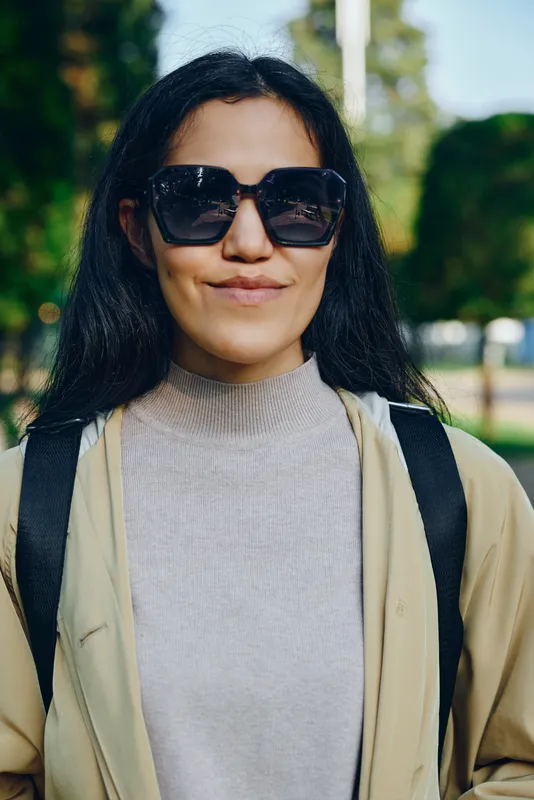 Sunglasses wearing woman in casual outdoor outfit stands in park, smiling, carrying backpack, urban vibe and sunny day, fashion conscious and relaxed exploration