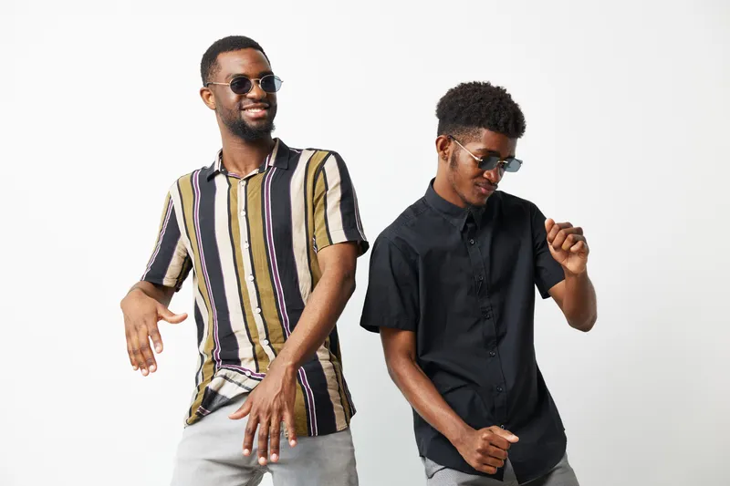 Two stylish black men dancing joyfully, wearing sunglasses and colorful shirts, exuding a playful and cheerful vibe in a bright setting.