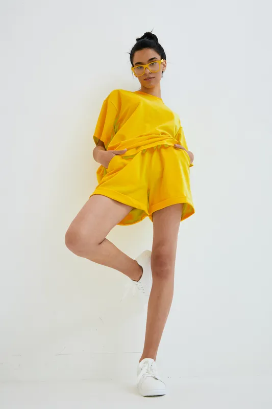 Young woman in bright yellow summer outfit posing confidently against white background, wearing matching shorts and top with casual white sneakers.