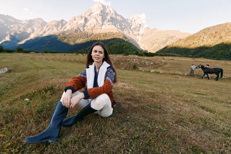 Woman sitting on meadow in mountains with horse in background, wearing boots and sweater, autumn landscape in open field, rural outdoor portrait and travel lifestyle.