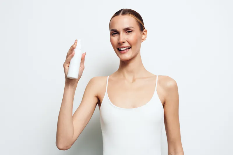 Young woman in a white tank top smiles while holding a bottle in a bright studio, showcasing a skincare product and a fresh radiant look.