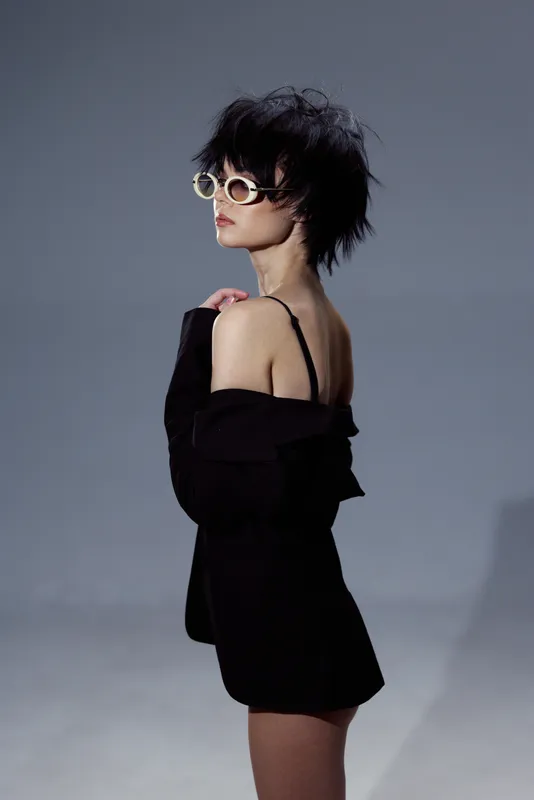 Stylish woman wearing sunglasses and black off-shoulder dress posing on gray background