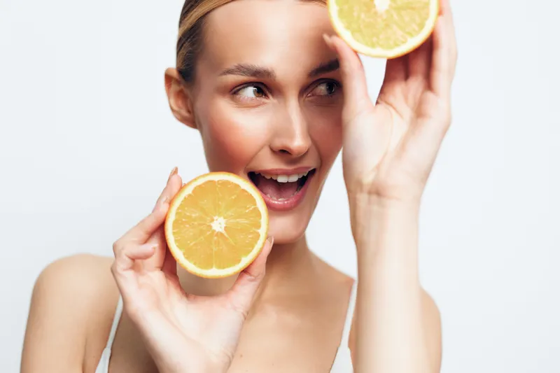 A happy young woman with clear skin holds two halves of a fresh orange near her face, smiling playfully against a plain white background. Concept for natural beauty and healthy lifestyle.