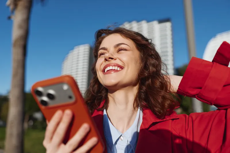 A smiling woman in a red jacket holds a smartphone and a gift, posing outdoors during a cheerful celebration with bright daylight and city buildings in the background